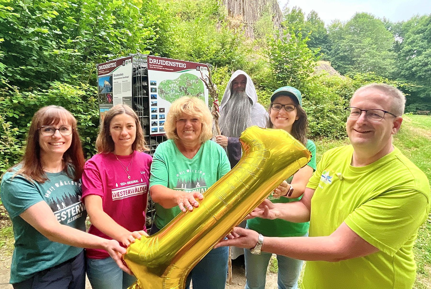 Six smiling people stand outdoors holding a shiny gold balloon shaped like the number one, with greenery and a Druidenstieg trail sign in the background. One person is dressed in a long robe and hood.